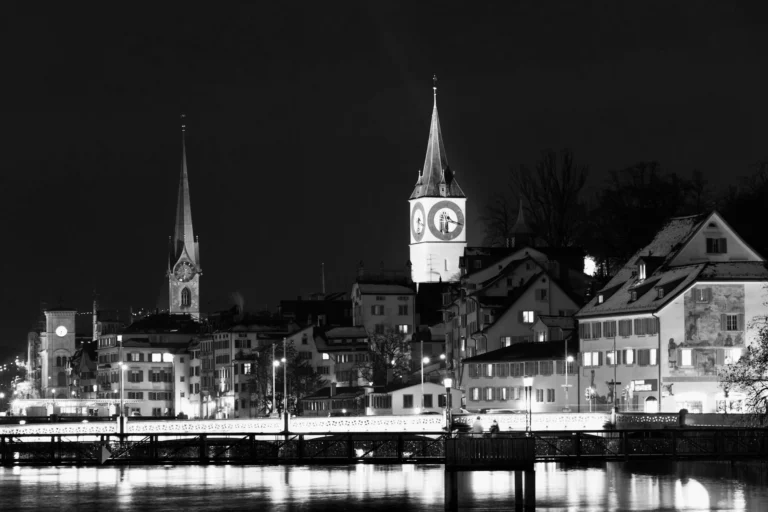 Black and white night view of Zurich with illuminated church towers and reflections on the river, symbolising the quiet rhythm of night work and recovery.